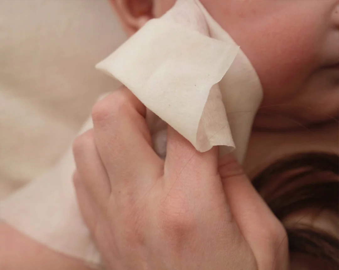 A close-up of a hand gently wiping a baby's cheek with a soft, white cloth.
