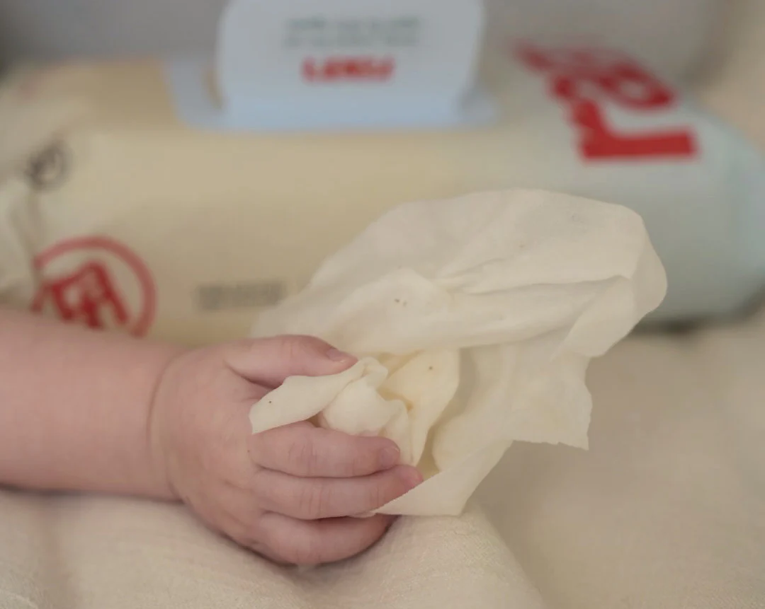 A close-up of a baby's hand holding a crumpled, off-white wipe on a light-colored surface.