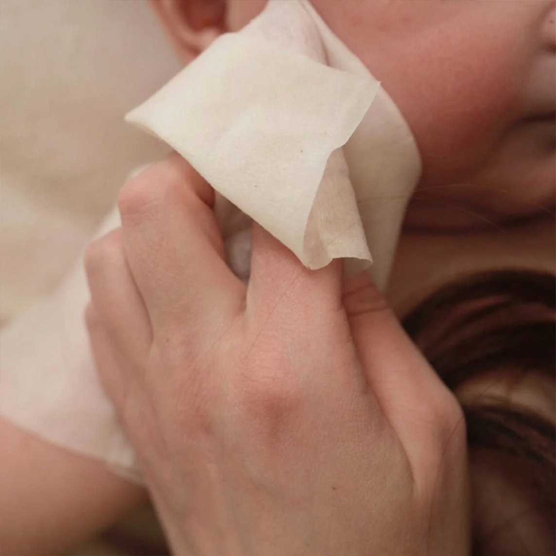 A close-up of a hand gently wiping a baby's cheek with a white cloth.