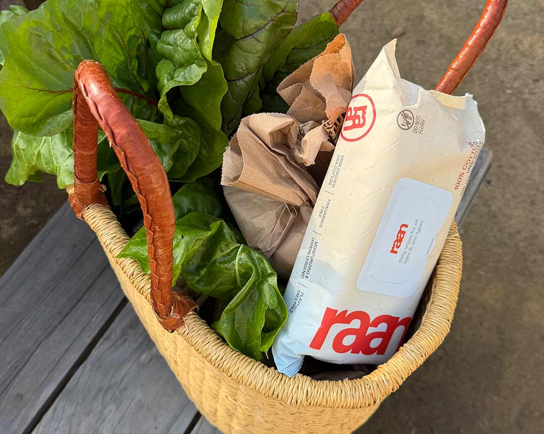 A close-up of a woven basket containing fresh leafy greens, a paper bag, and a package of wipes.