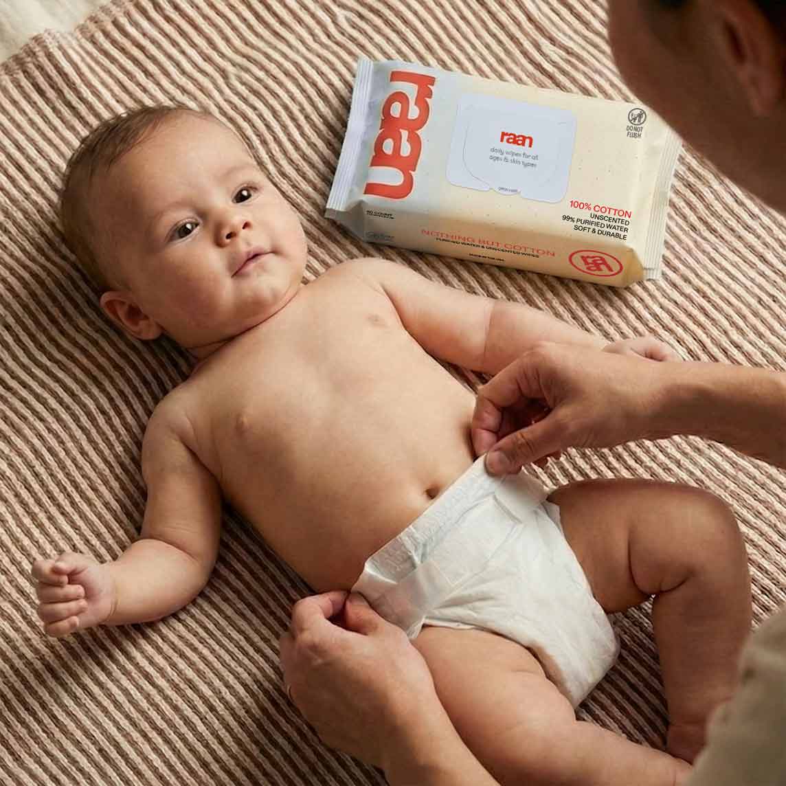 A baby lies on a striped blanket while an adult changes their diaper, with baby wipes nearby.