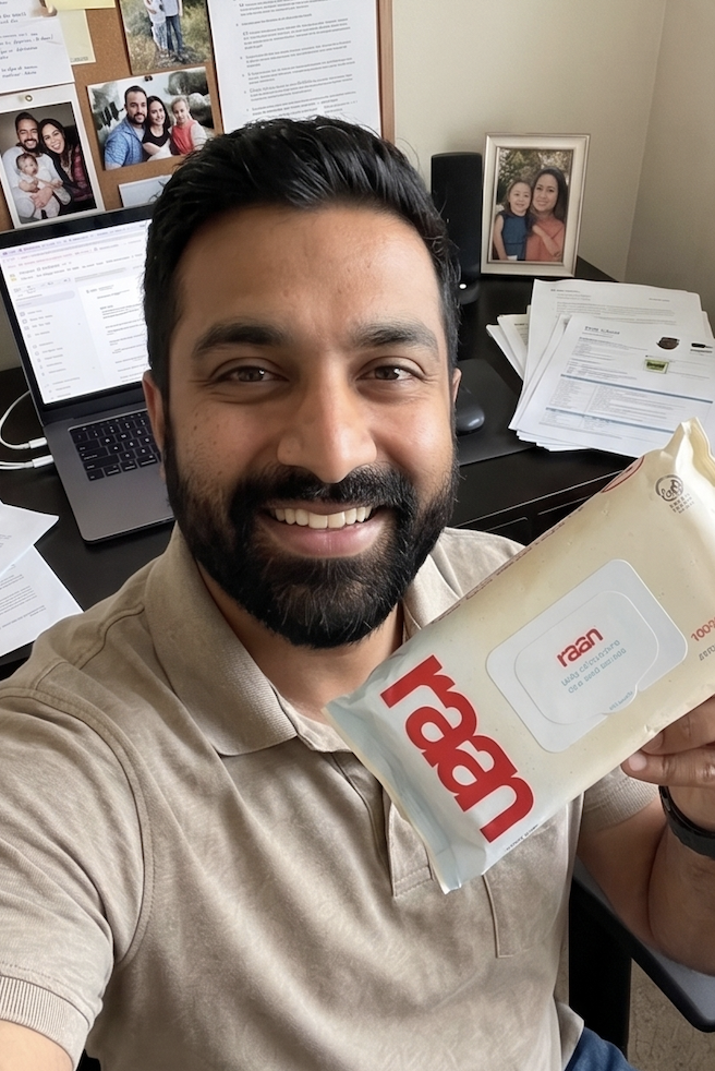 A smiling man with a beard sits at a desk holding a package of Raan brand wipes.