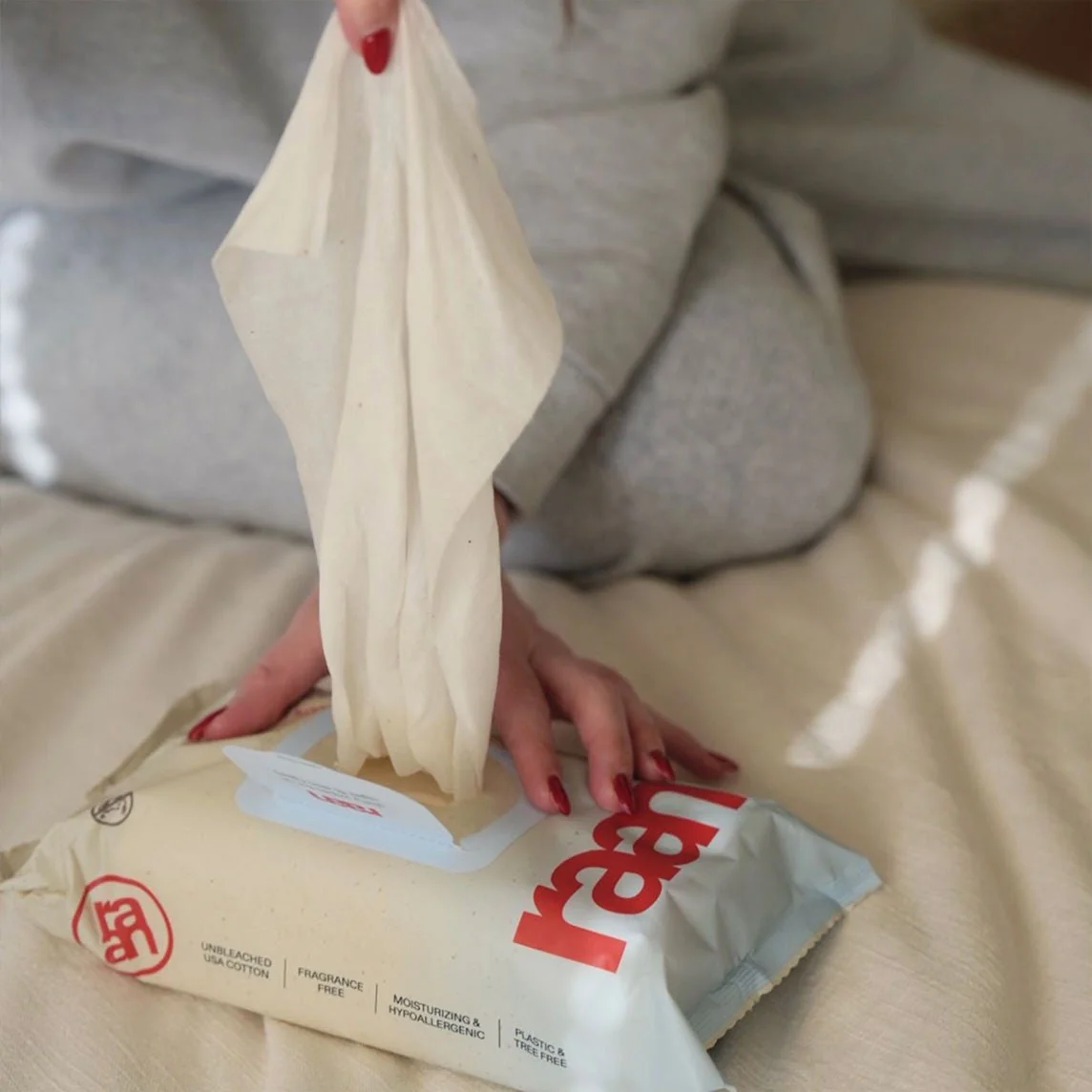 A person with red nails pulls a wipe from a package of Rael brand wipes on a bed.