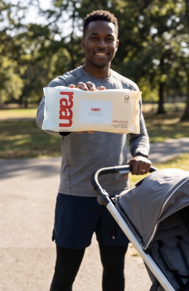 A smiling man holds a package of raan wipes while pushing a baby stroller in a park.