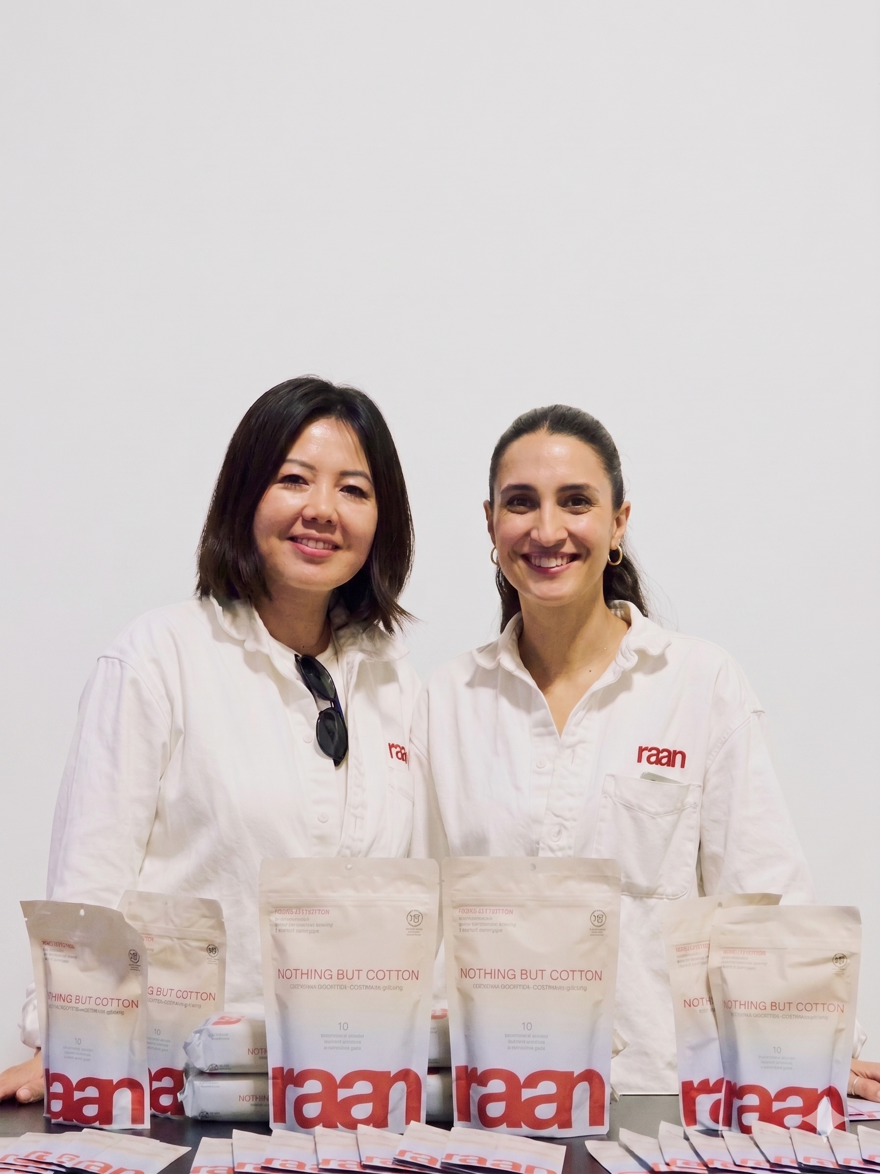 Two women in white shirts smile behind a table displaying packages of 'Nothing But Cotton' by Raan.