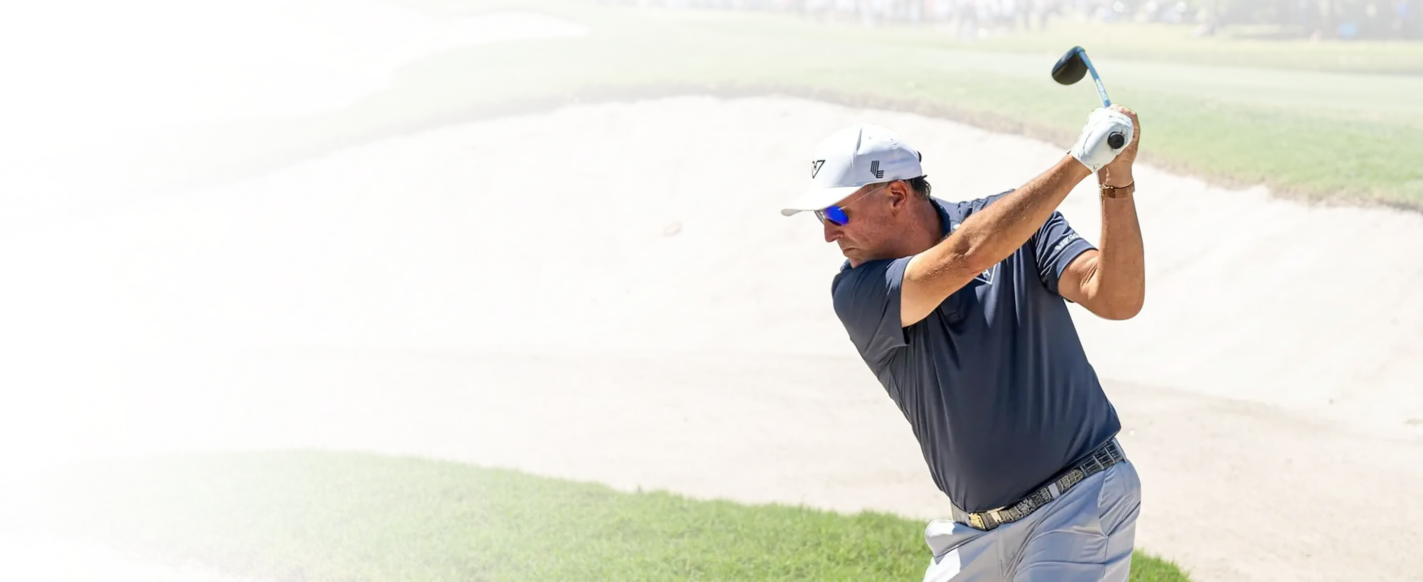A male golfer in a white cap and sunglasses mid-swing in a sand bunker on a golf course.