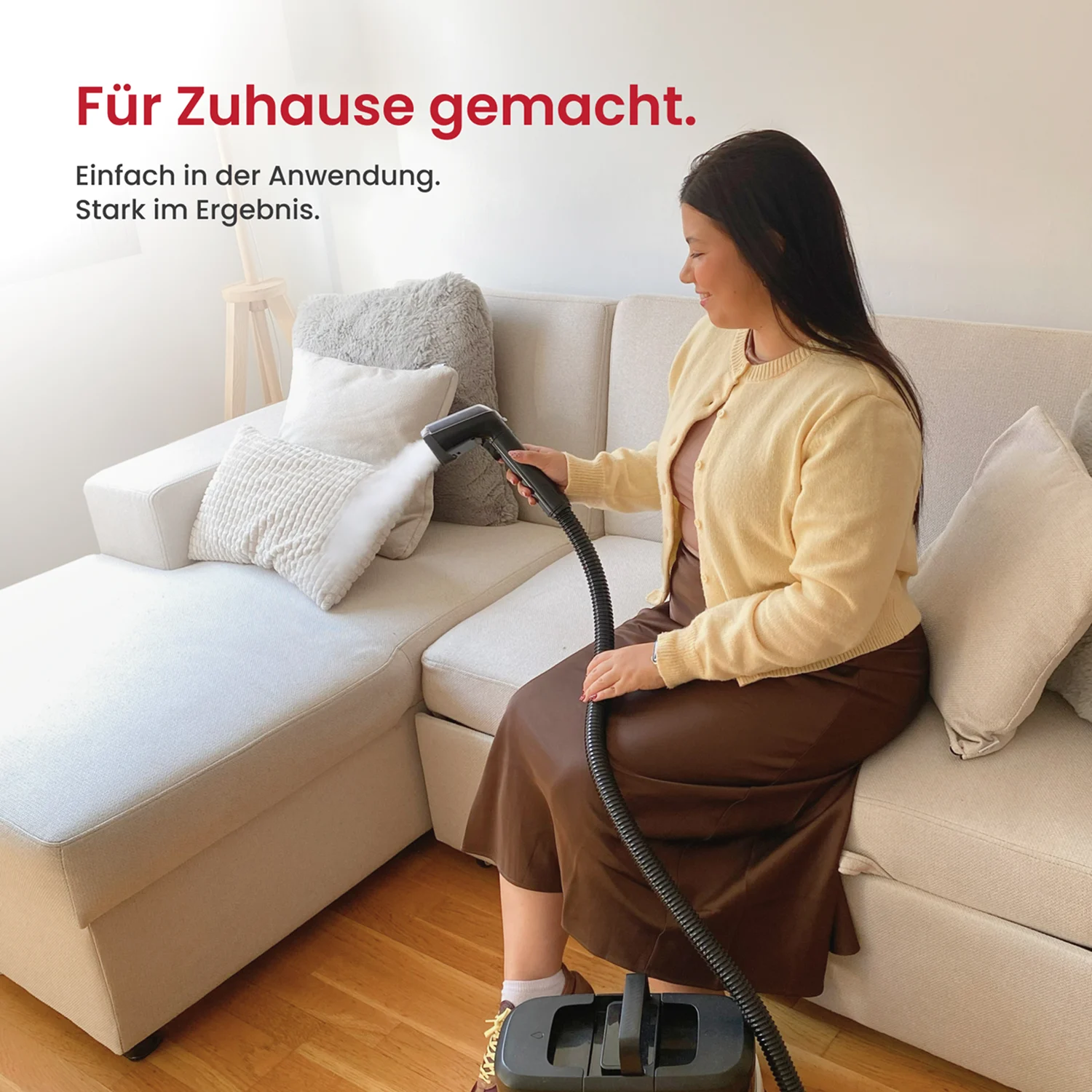 A woman sits on a light-colored sofa while using a steam cleaner on a pillow.