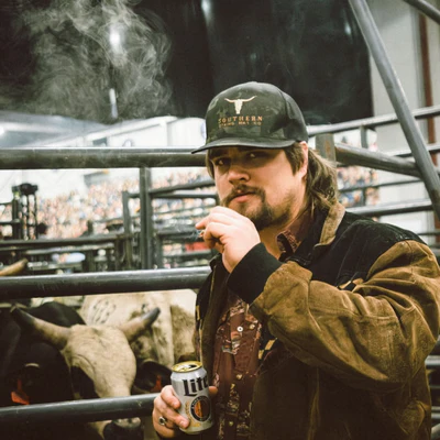 Man in a cap smoking and holding a drink near a bull pen.