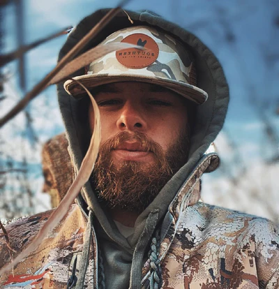 Person in camouflage clothing and hat outdoors, with a blurred background.