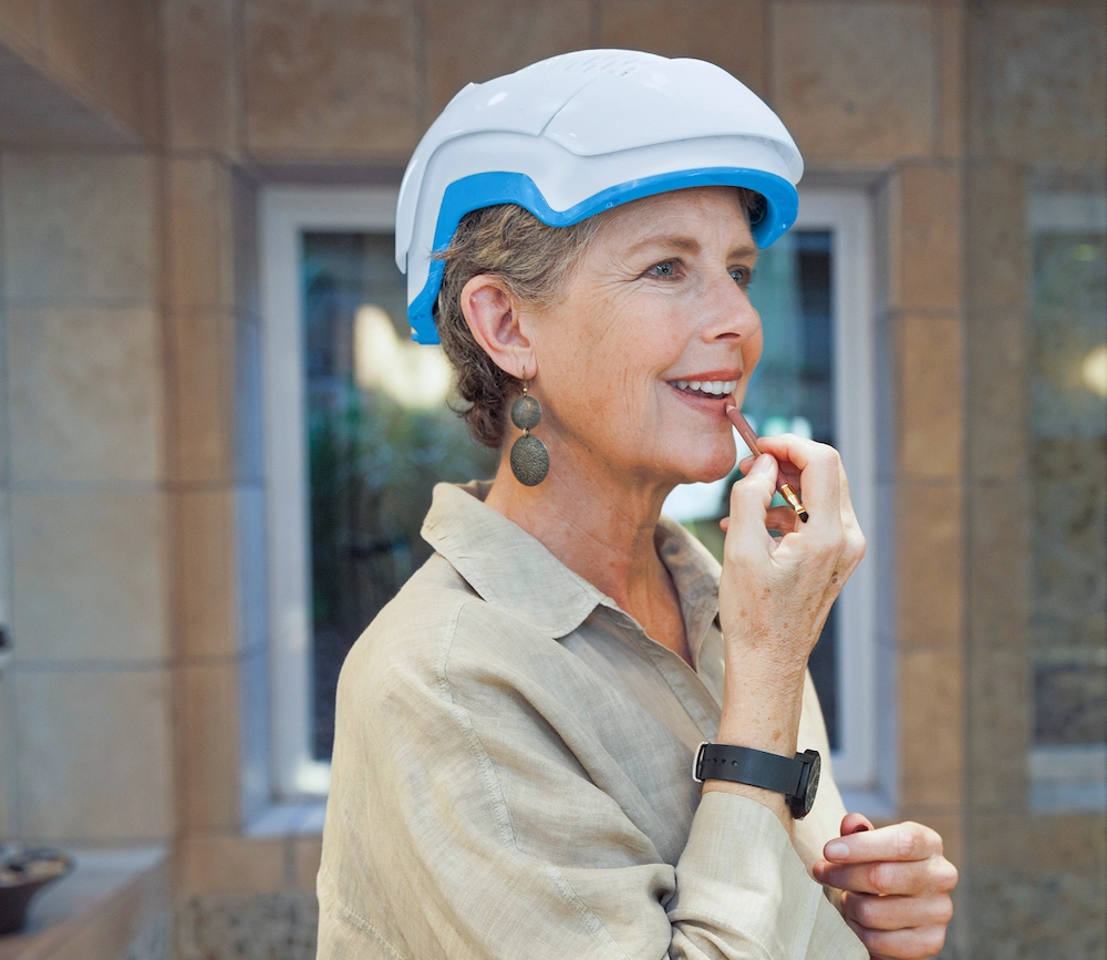 A person applying lipstick while wearing a white helmet indoors.