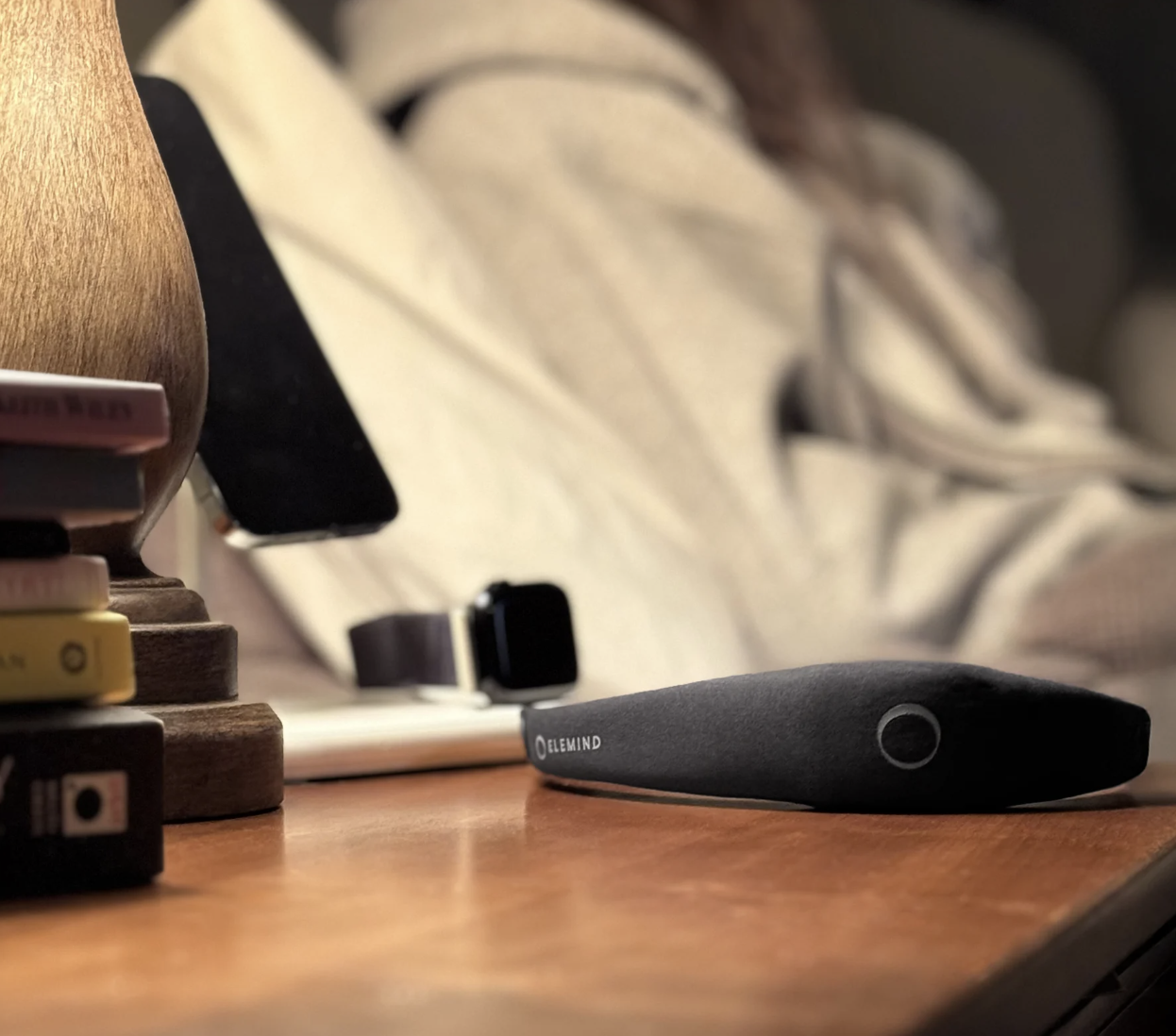 A black Elemind headband on a nightstand next to a lamp, charging phone, and watch, with a person in bed.