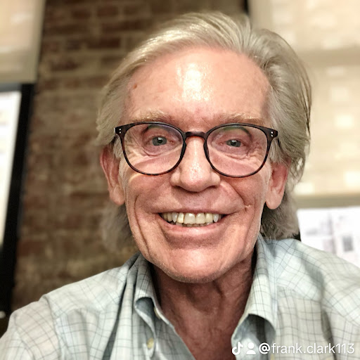 A smiling older man with gray hair and glasses, wearing a light-colored plaid shirt.