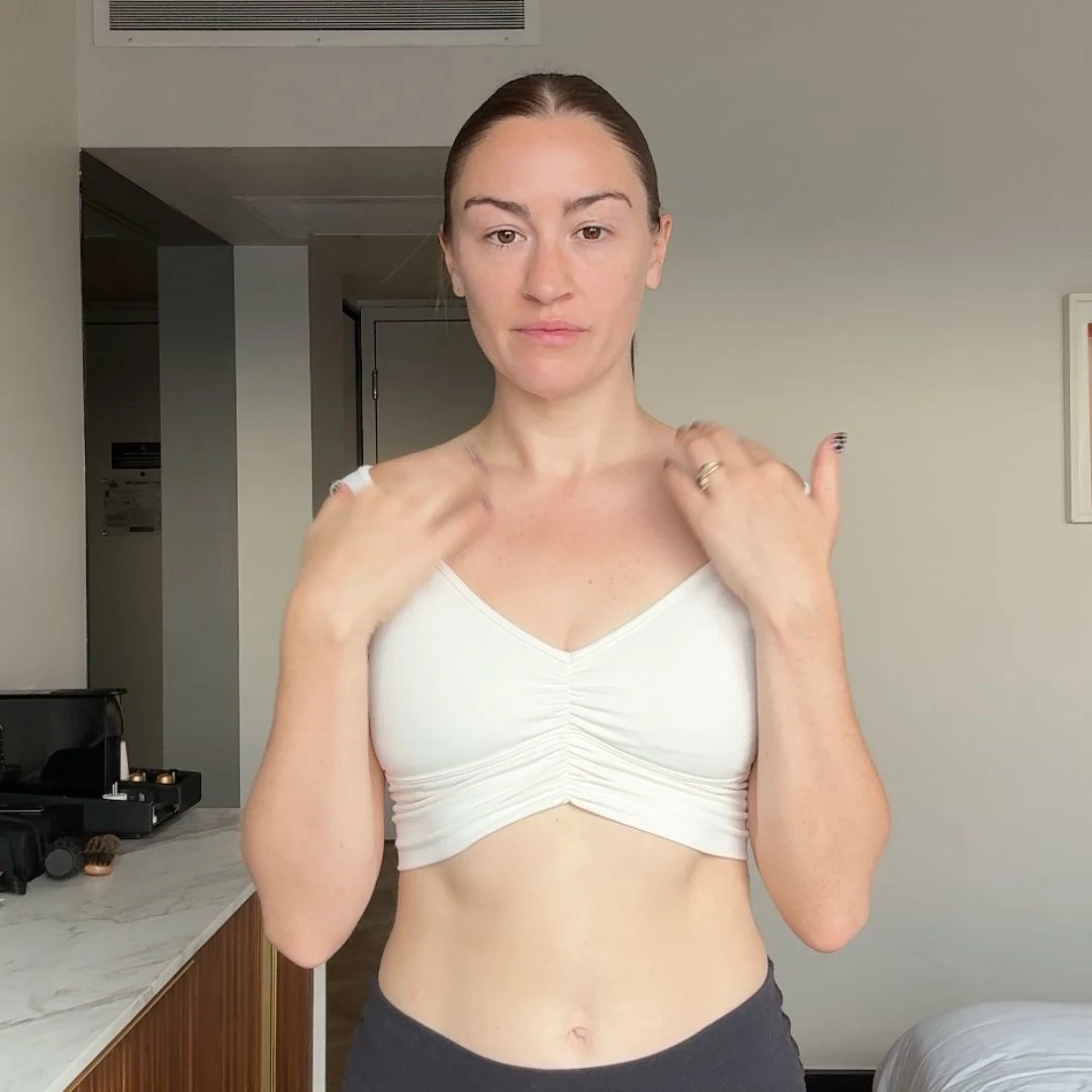 A woman with her hair pulled back stands in a room wearing a white crop top and black leggings.