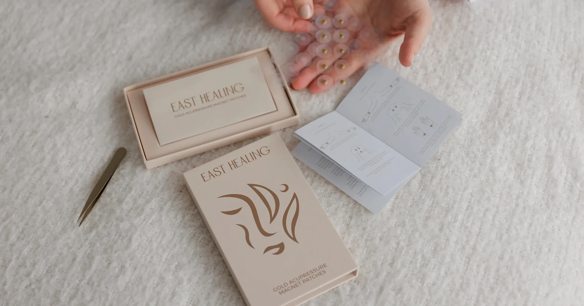 A person unboxes East Healing gold acupressure magnet patches with an instruction manual and tweezers nearby.
