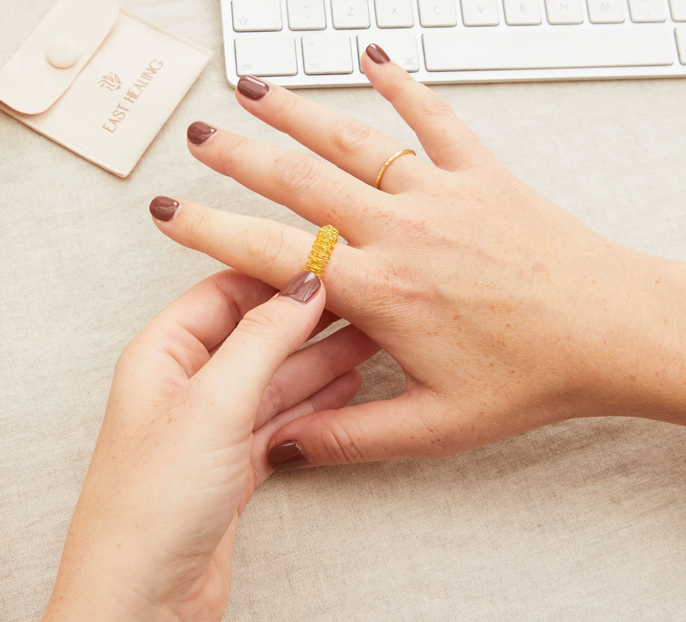 A person with brown nail polish slides a gold, coiled wire ring onto their finger.