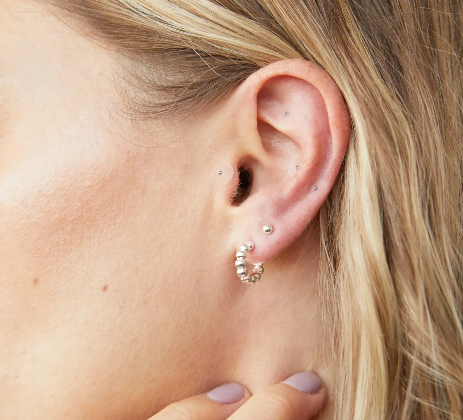 A close-up of a woman's ear with multiple silver piercings, including a beaded hoop and several studs.