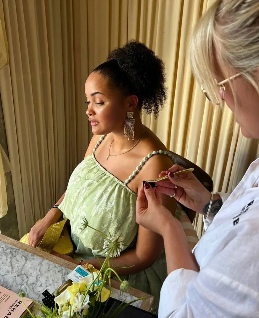 A woman in a light green top has an adornment applied to her shoulder by another woman with tweezers.