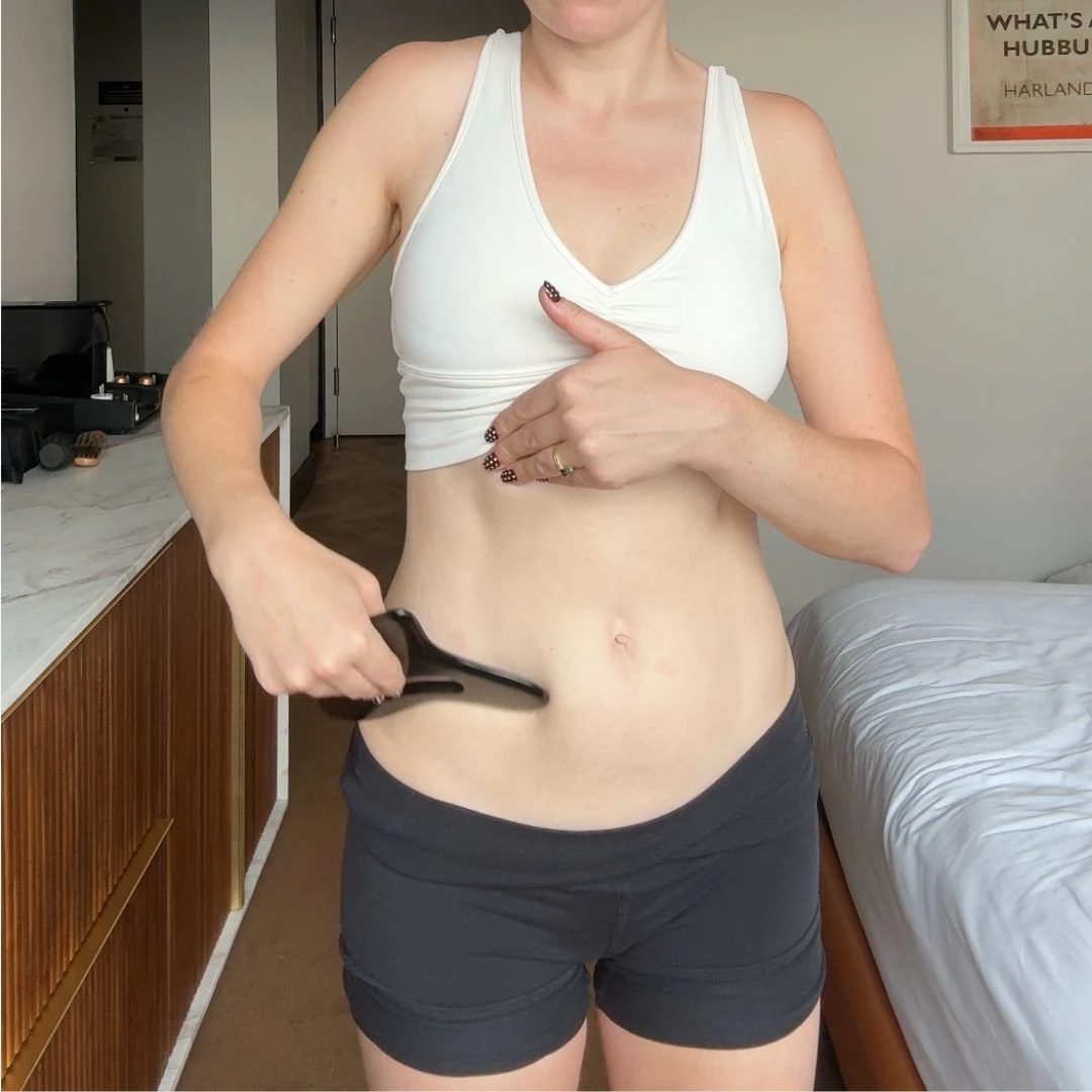 A woman in a white sports bra and black shorts uses a black massage tool on her stomach.