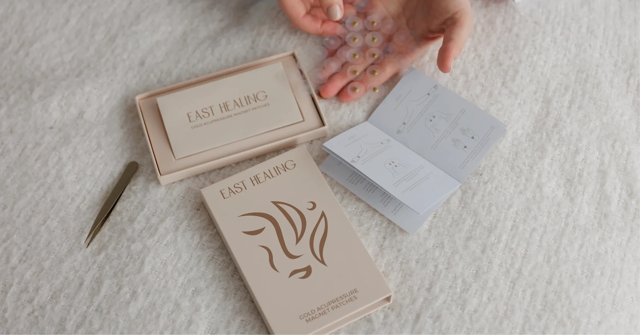 A person unboxing East Healing gold acupressure magnet patches with tweezers and an instruction manual on a white blanket.