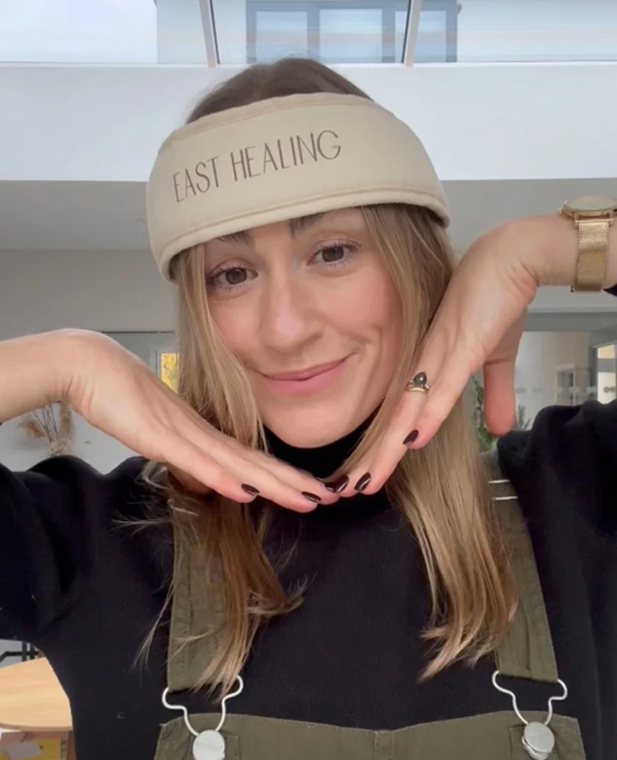 A woman wearing a headband that says 'EAST HEALING' smiles while framing her face with her hands.