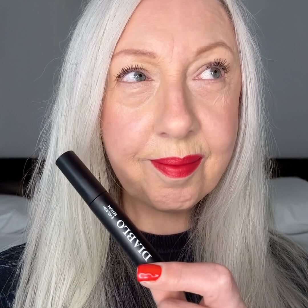 A woman with silver hair and red lipstick holds a black tube of Diablo Eyelash Serum.