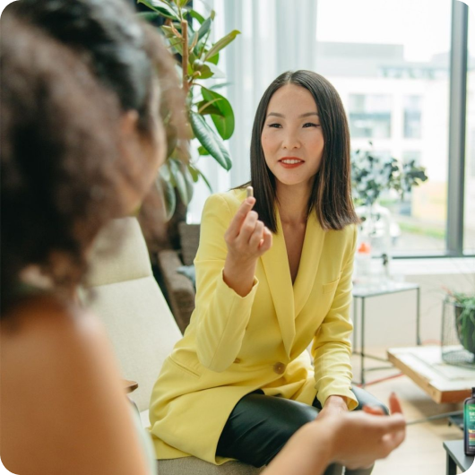 A woman in a yellow blazer talking with another person in an office setting.