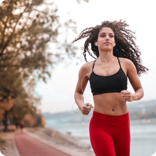 Woman jogging on a tree-lined path by the water, wearing earphones.