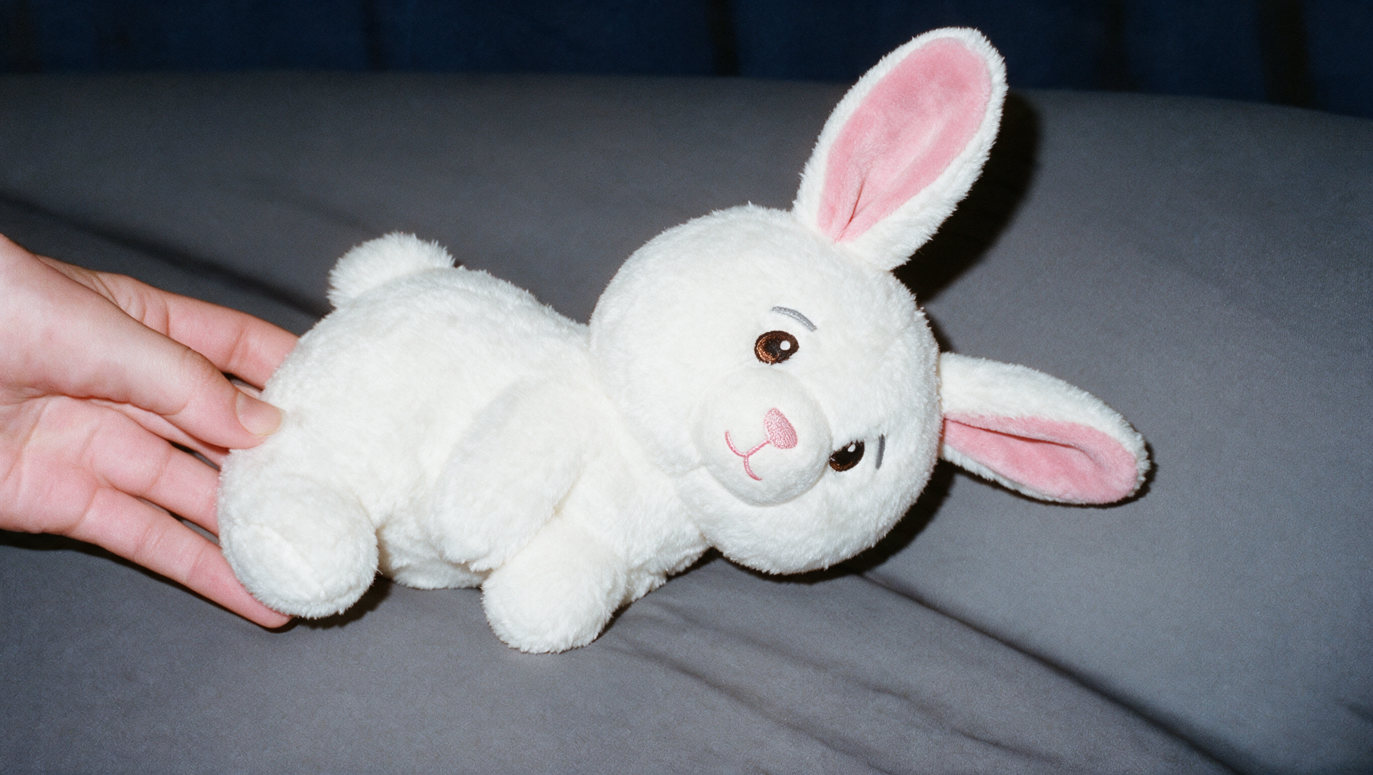 A person's hand holds a white stuffed rabbit with pink ears on a gray surface.