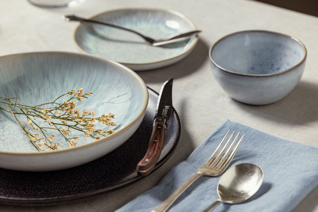 A rustic table setting with blue ceramic dinnerware, silverware, and a sprig of dried flowers in a bowl.