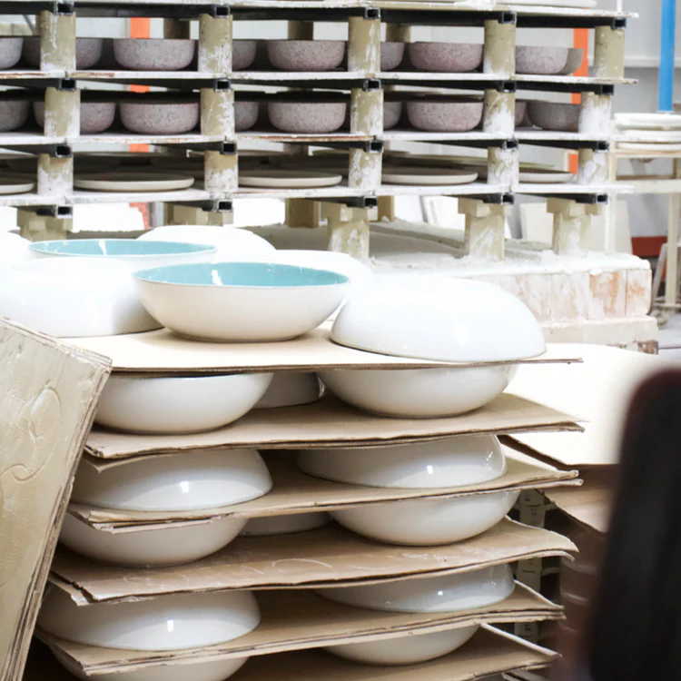 White and turquoise ceramic bowls sit on shelves in a pottery workshop, waiting to be fired.