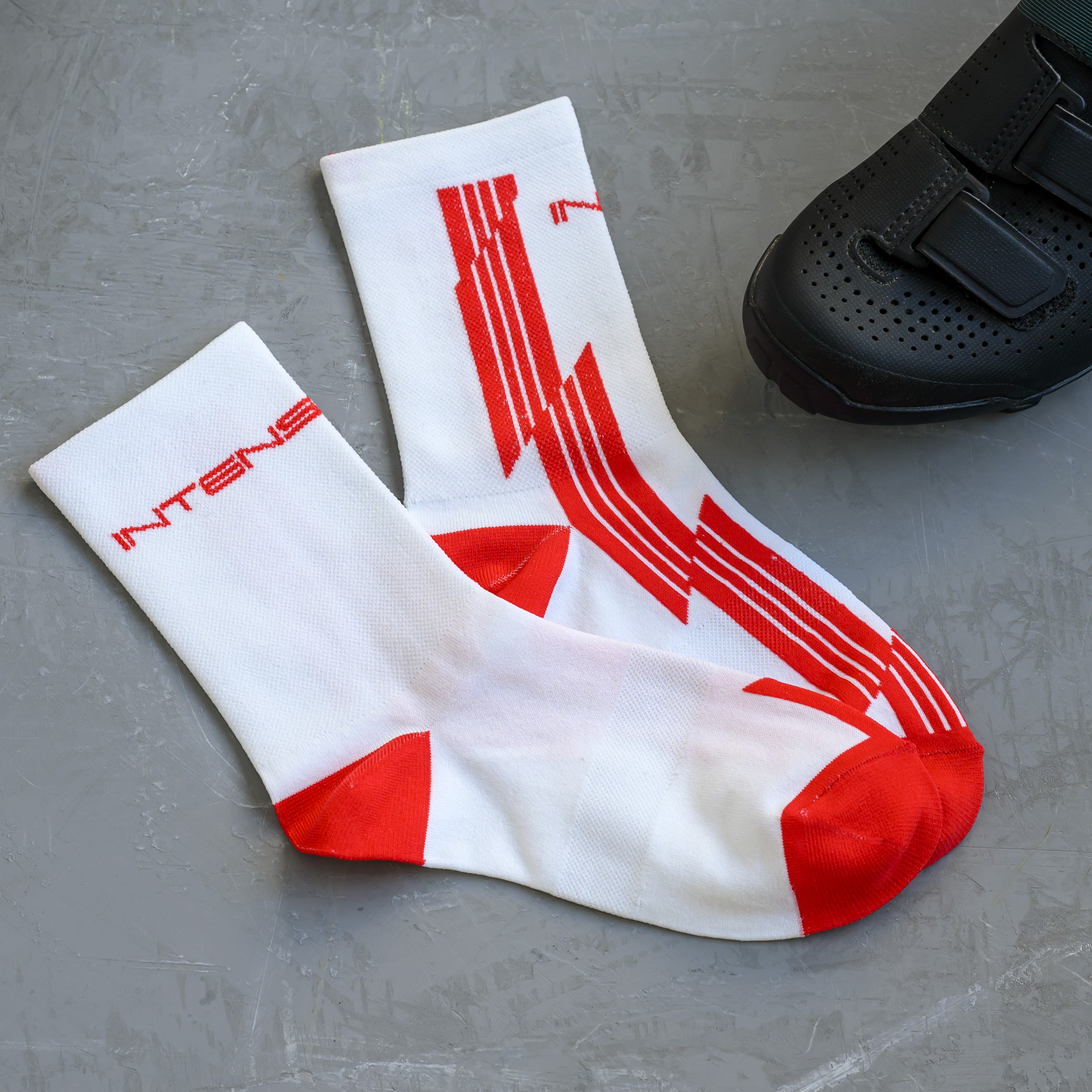 A pair of white and red Intense brand cycling socks next to a black cycling shoe on a grey surface.