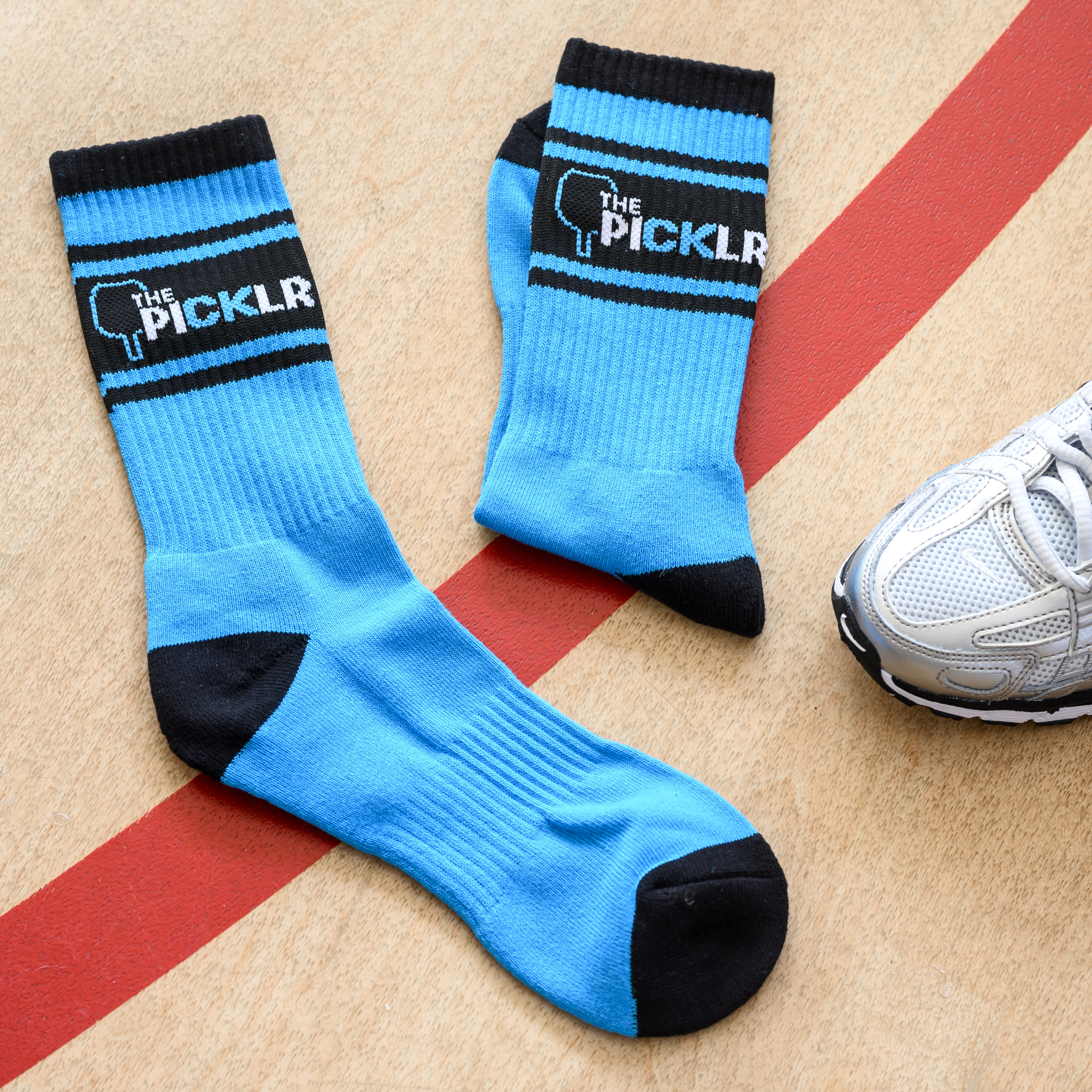 A pair of blue and black 'The Picklr' athletic socks on a court next to a sneaker.