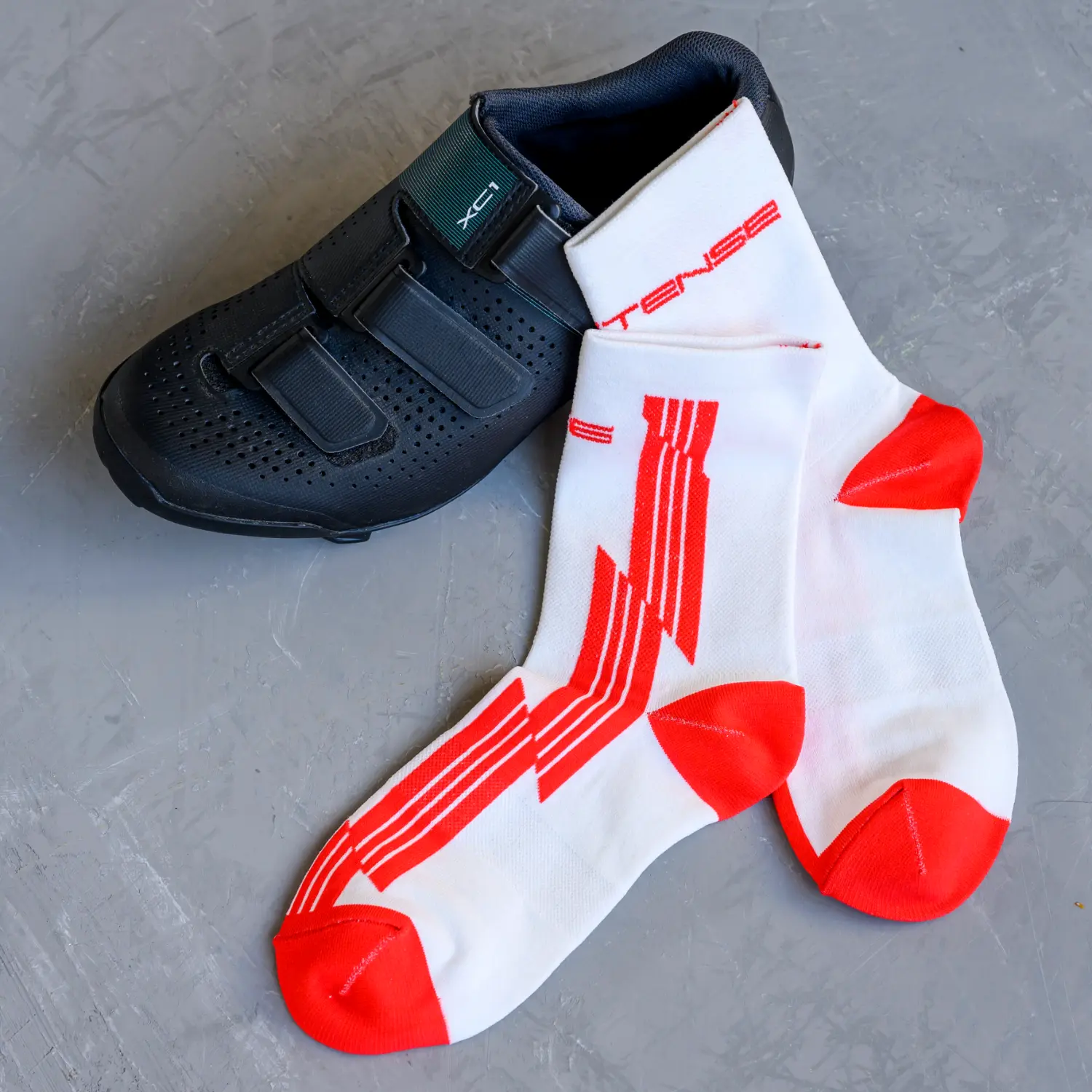 A single black cycling shoe lies next to a pair of white and red athletic socks on a gray surface.