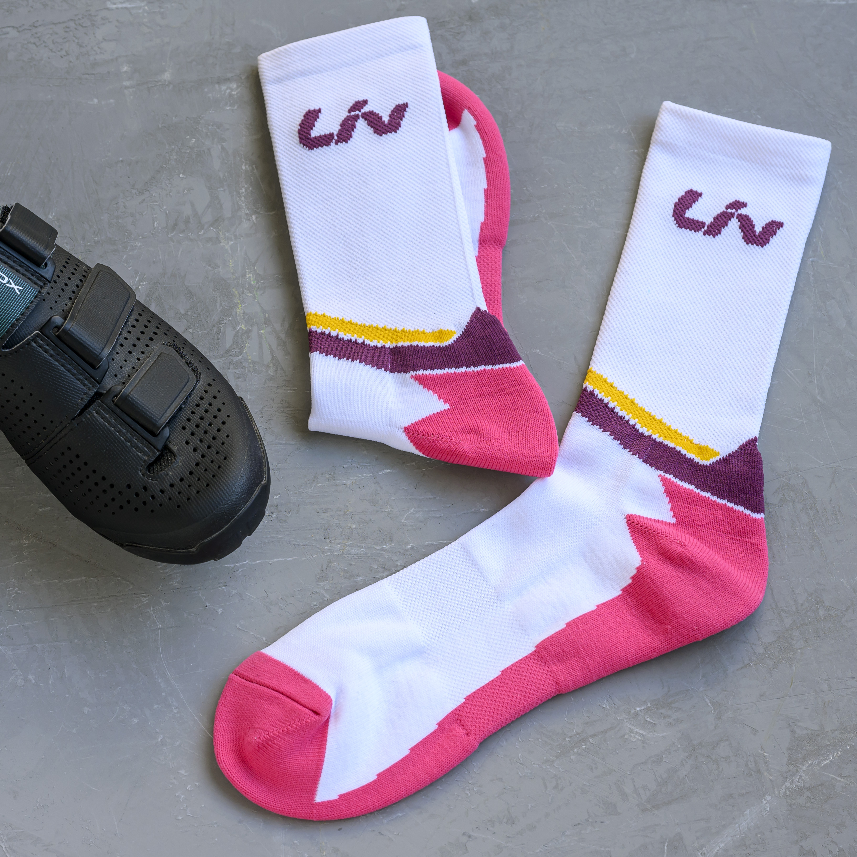 A pair of white cycling socks with pink, purple, and yellow details next to a black cycling shoe.