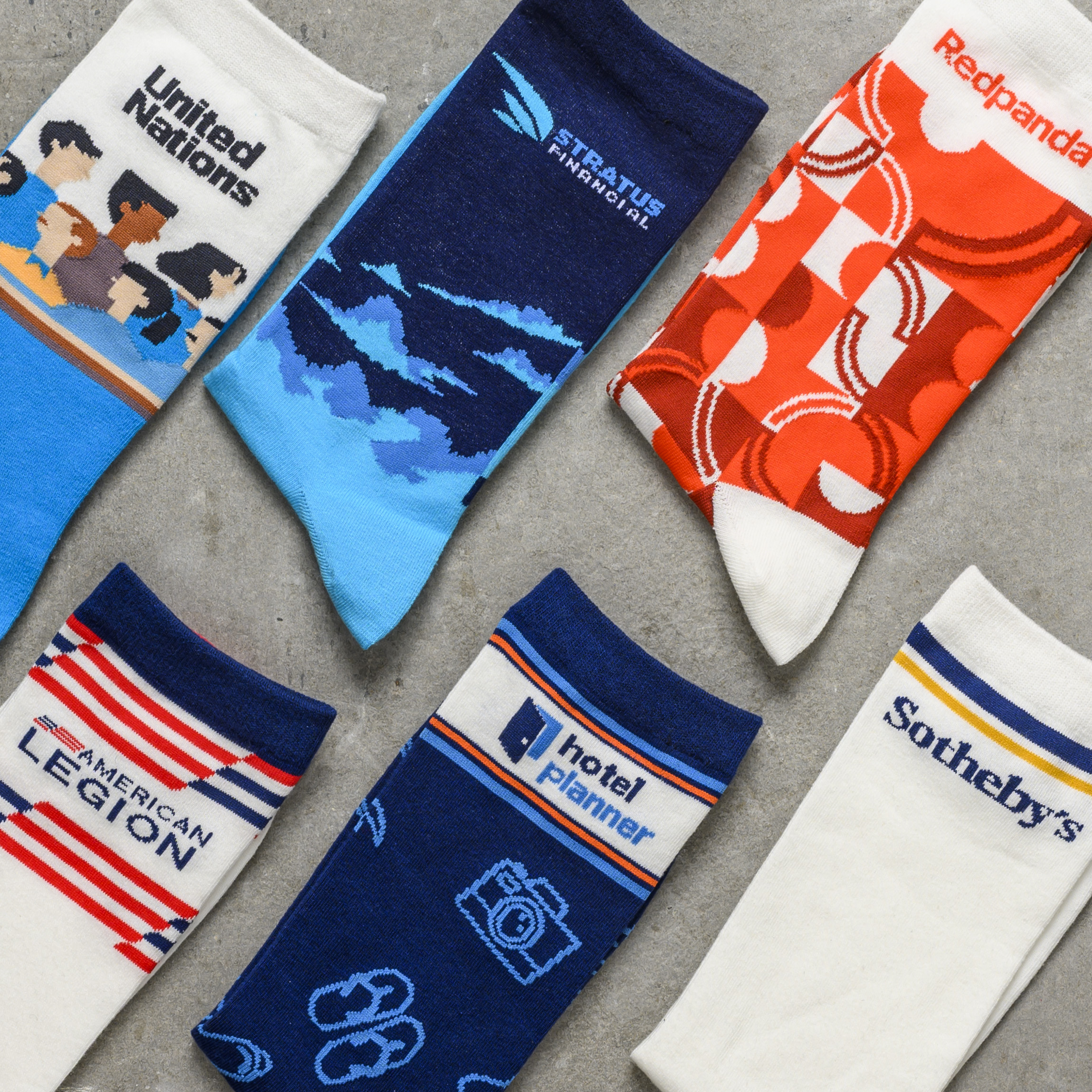 An overhead view of six different pairs of custom-designed promotional socks arranged on a gray concrete surface.