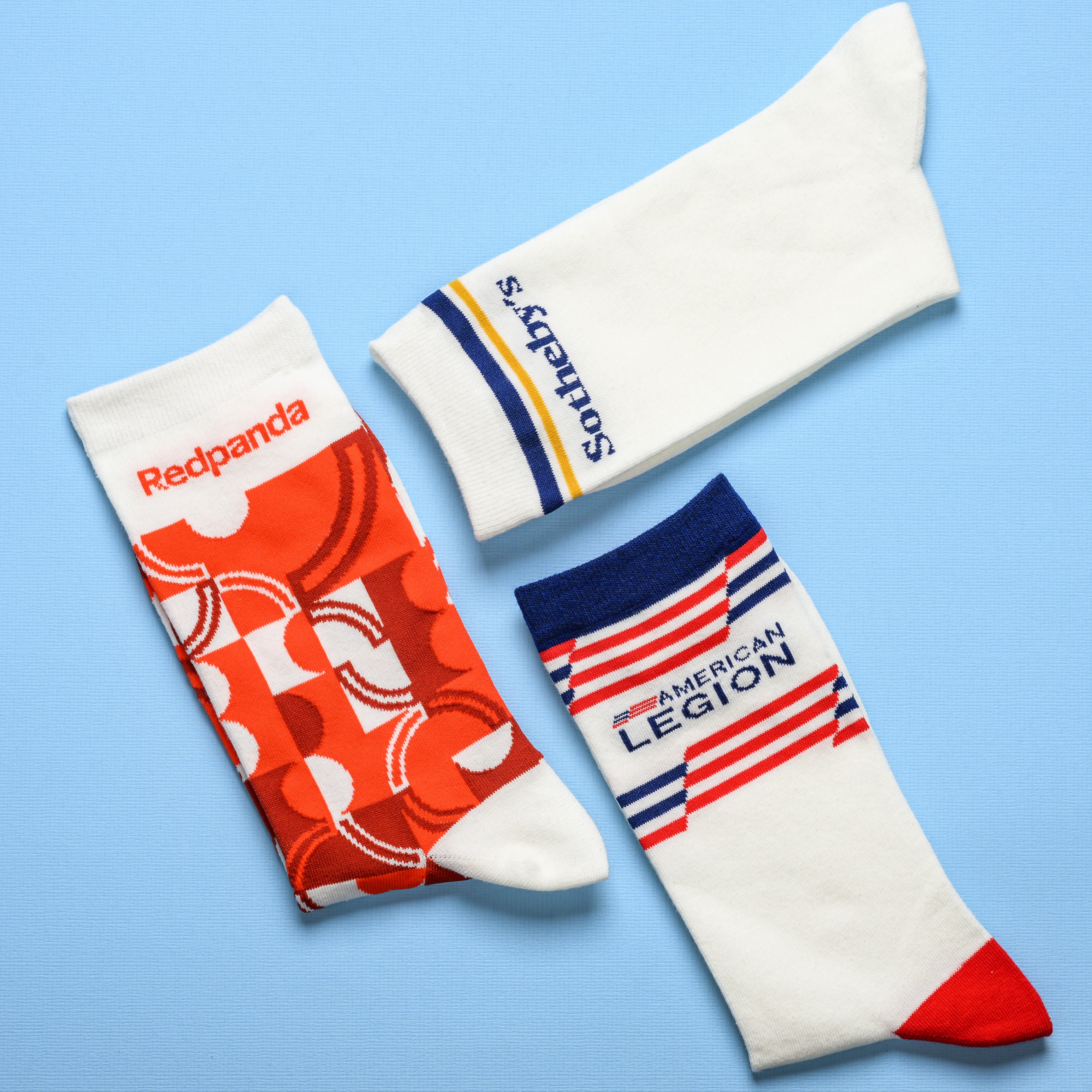 An overhead view of three different custom branded socks from Redpanda, Sotheby's, and American Legion on a blue background.