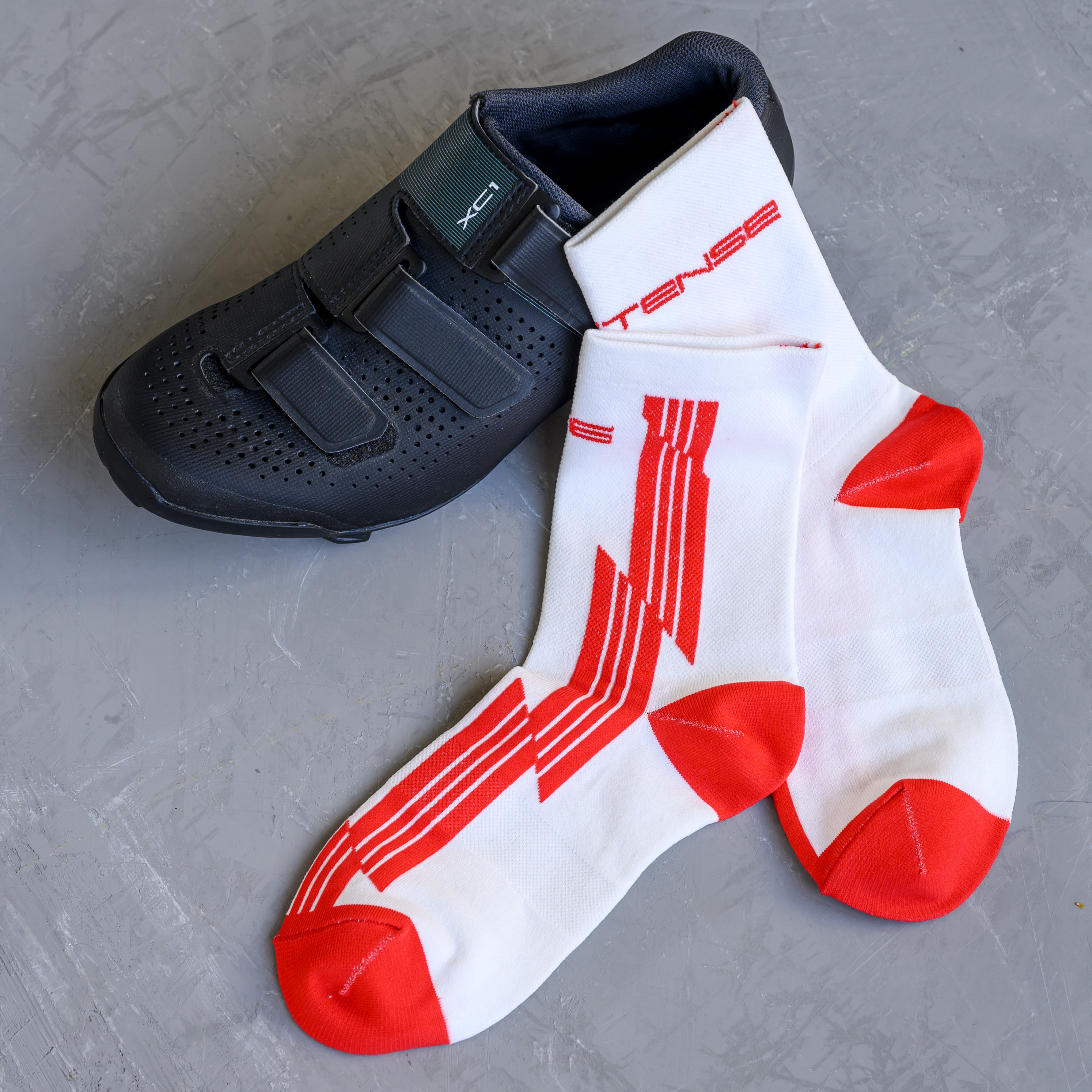 A single black cycling shoe lies next to a pair of white and red athletic socks on a gray surface.