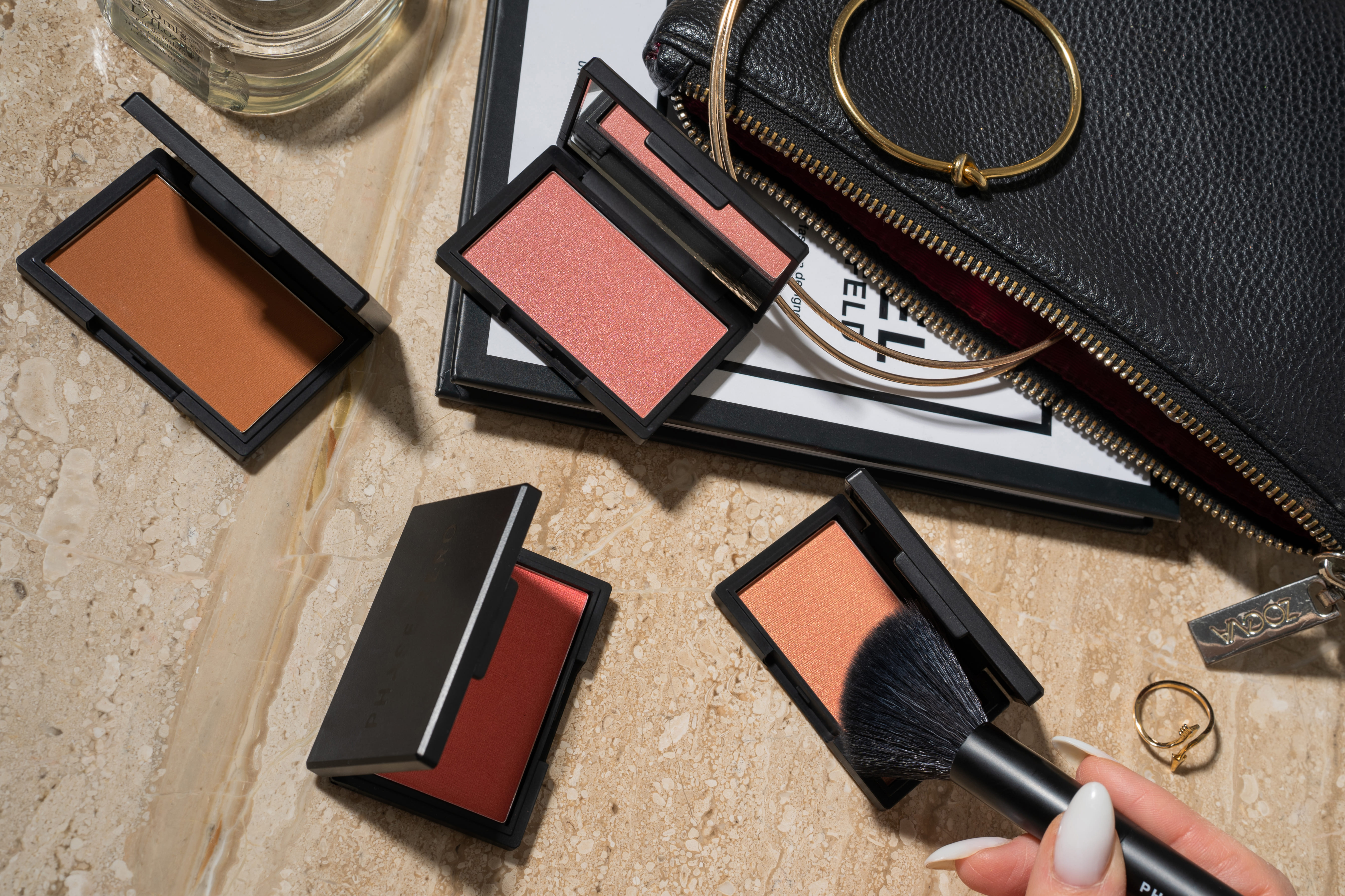 Makeup brushes and blushes arranged on a marble surface with a black bag.