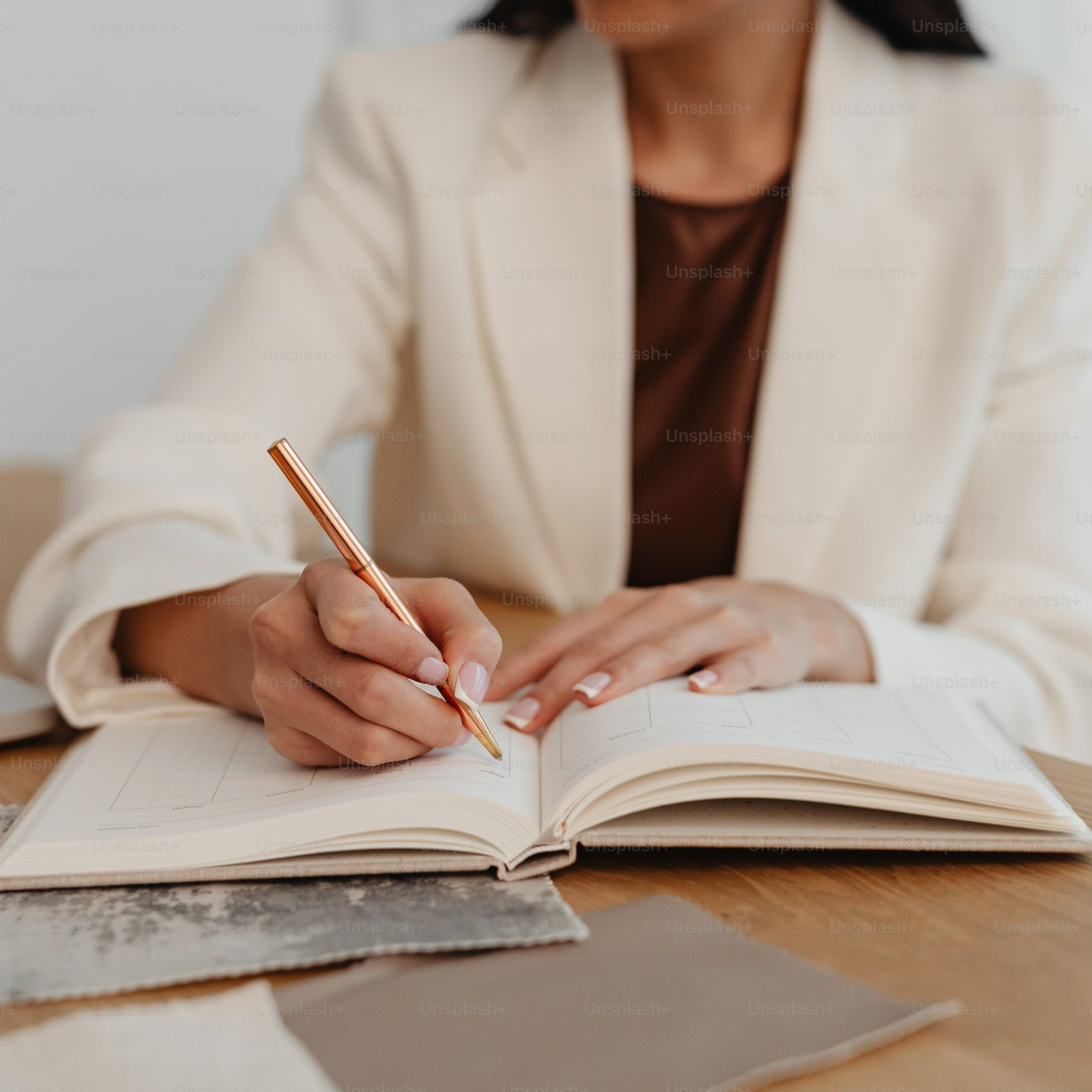 A person in a cream blazer writes with a gold pen in an open planner on a desk.
