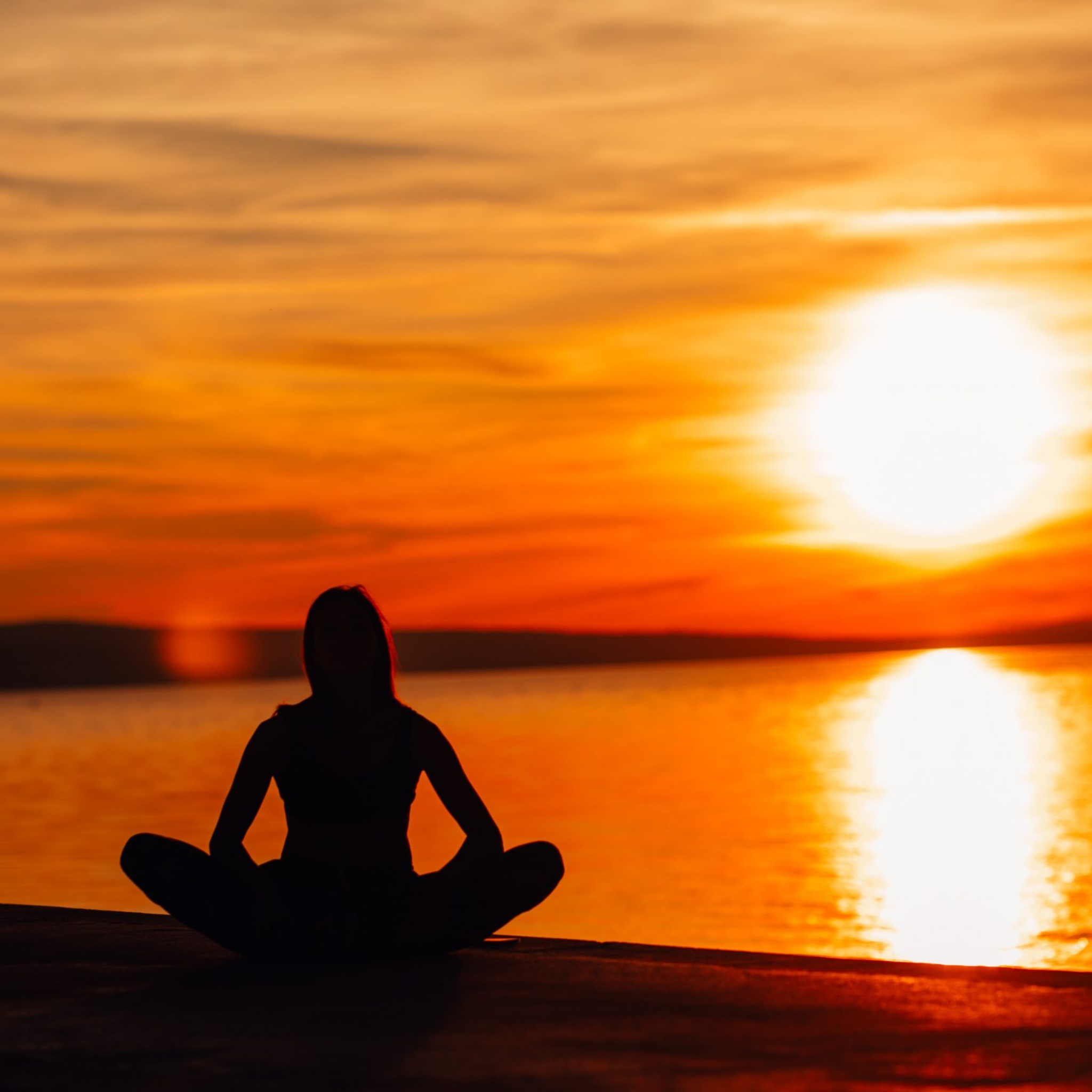 A silhouette of a person meditating on a dock, facing a vibrant orange sunset over the water.
