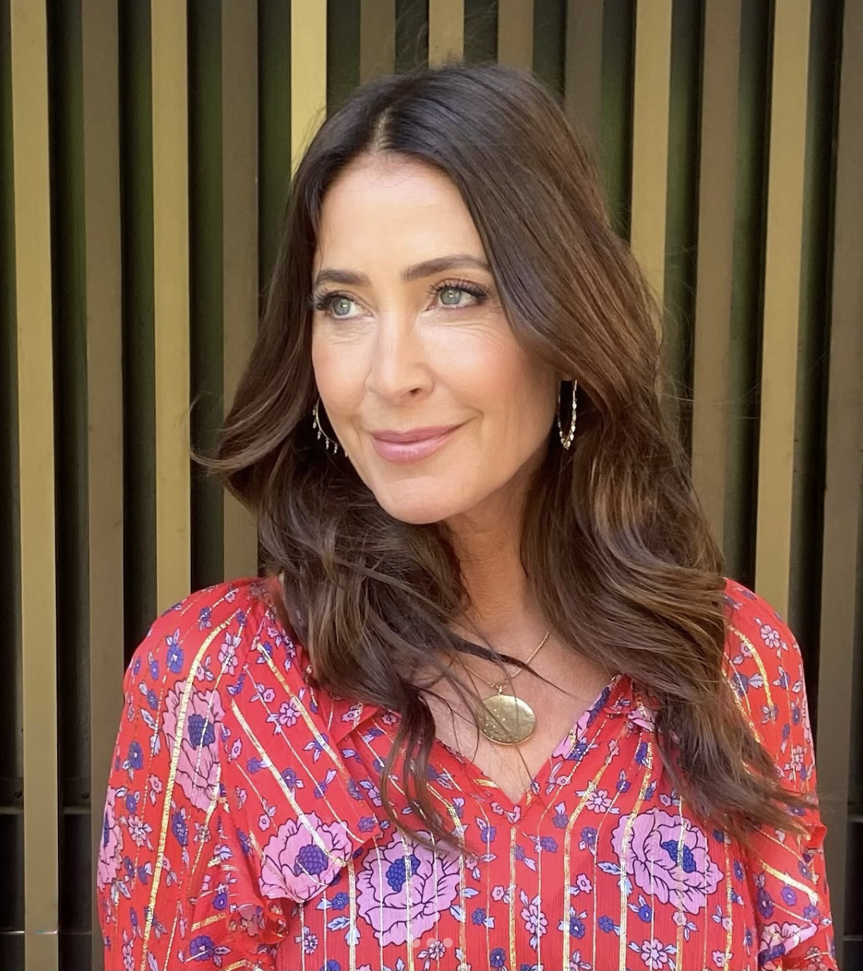 Woman with long brown hair in a floral red dress, standing against a striped background.