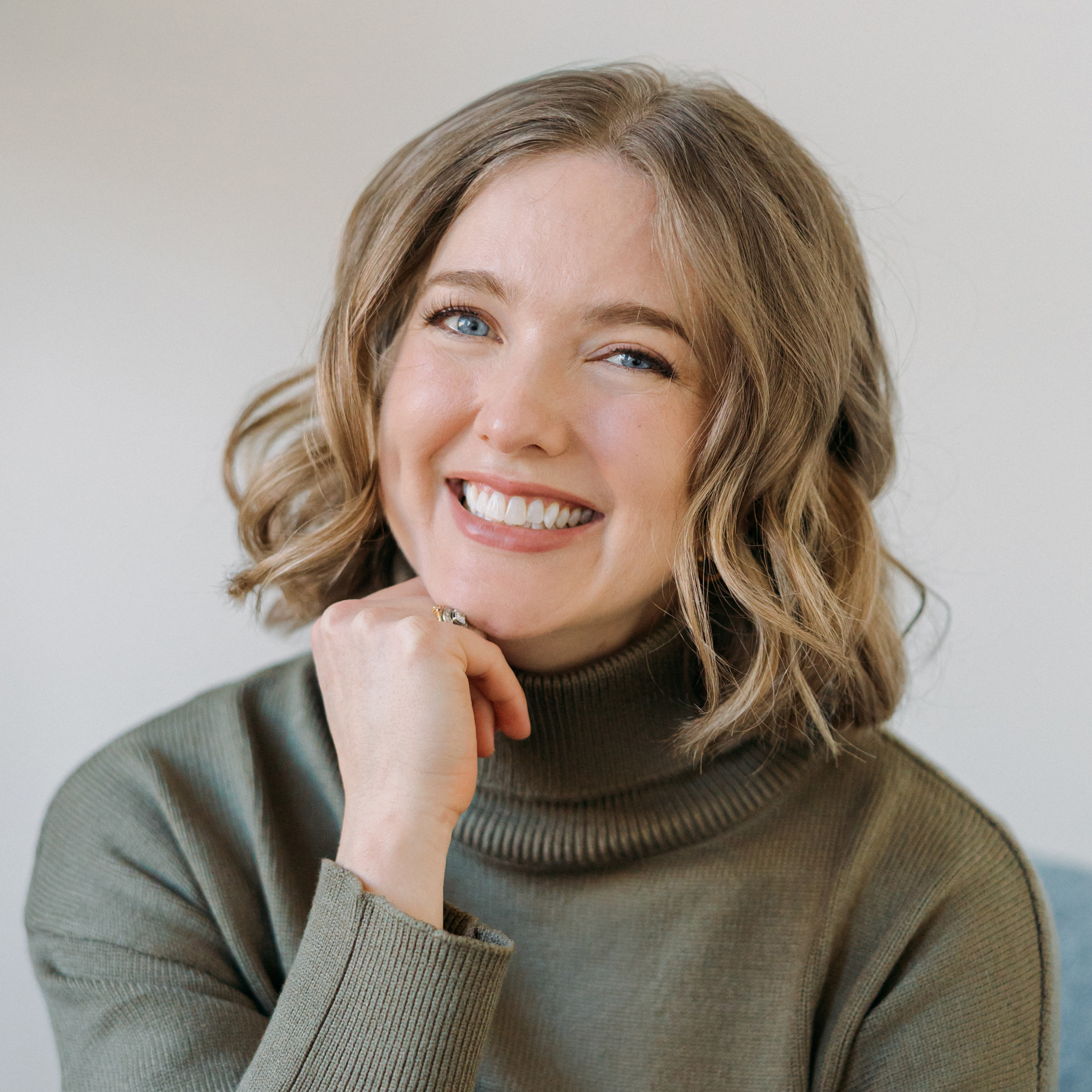 A smiling woman with blue eyes and wavy blonde hair wearing a green turtleneck sweater, resting her chin on her hand.