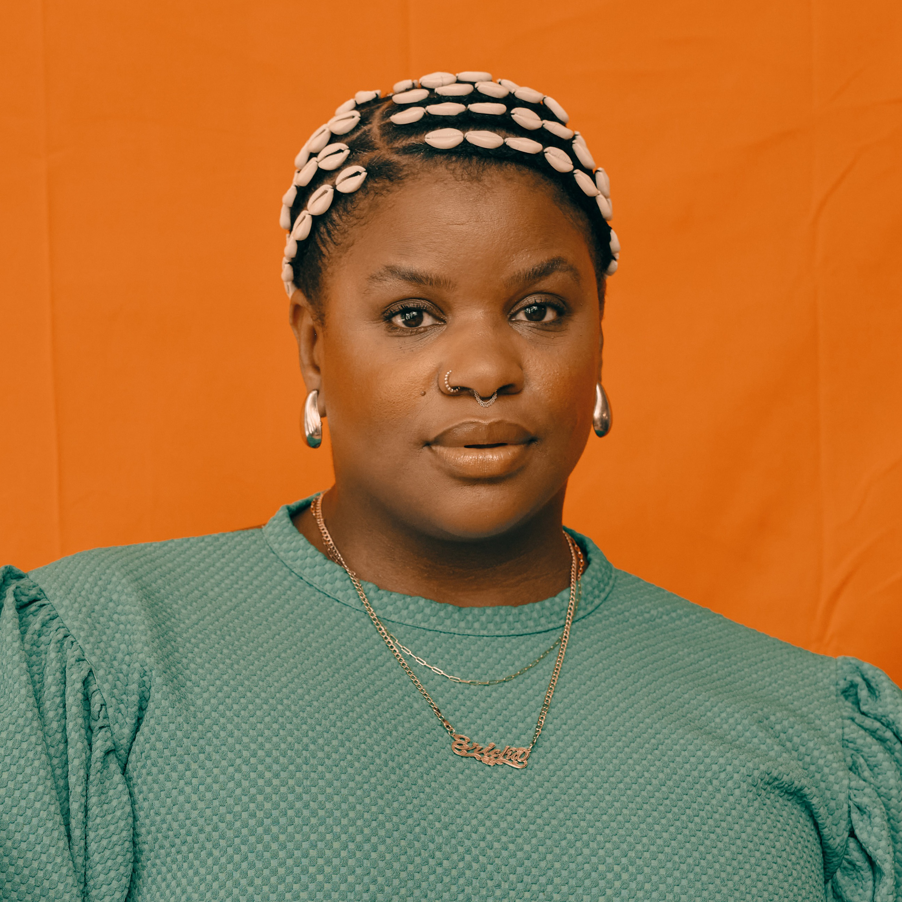 A portrait of a Black woman with cowrie shells in her braided hair against a solid orange background.