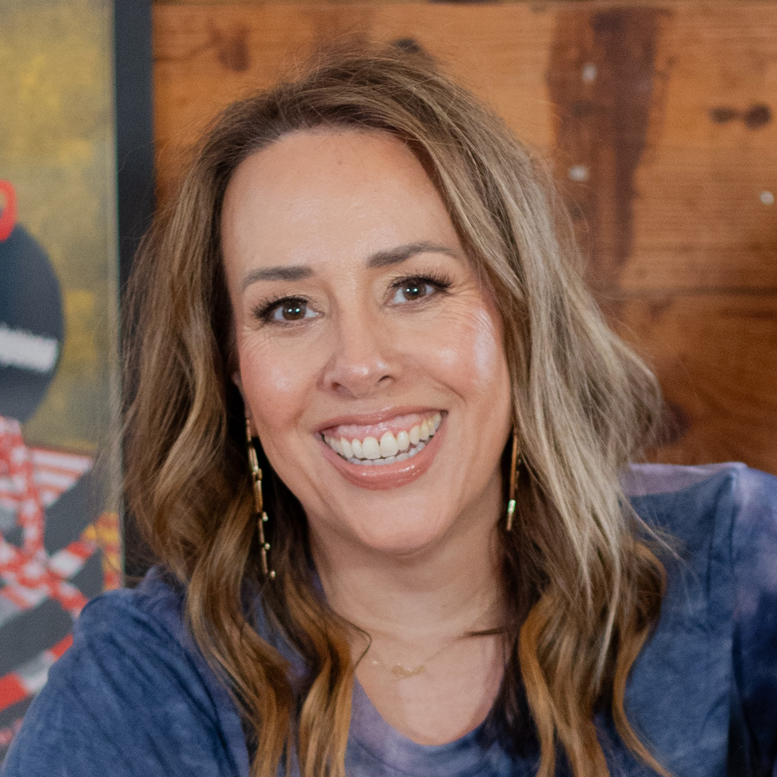 A headshot of a smiling woman with highlighted brown hair, wearing a blue shirt and long gold earrings.