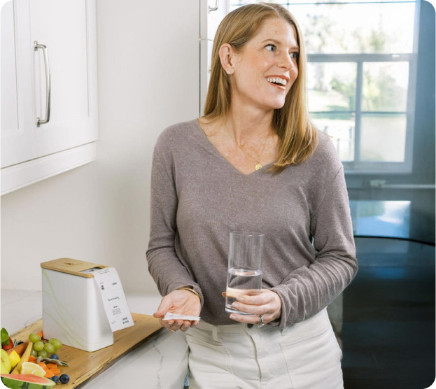 Woman in a kitchen holding a glass of water and a small object, smiling.