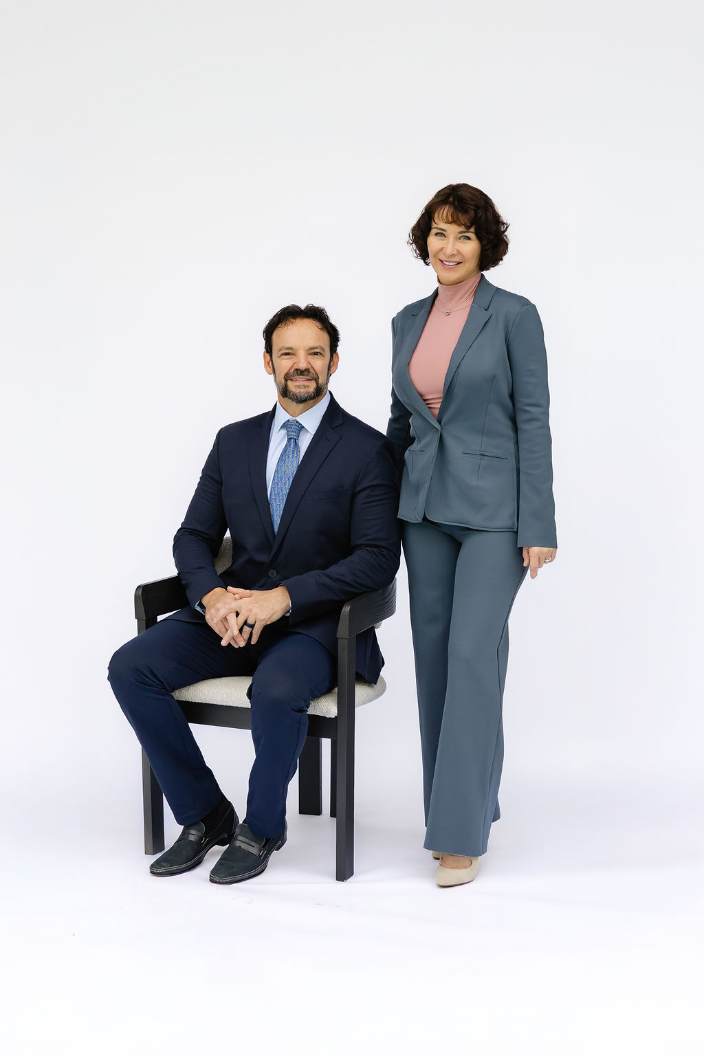 A man in a suit sits in a chair next to a woman in a pantsuit against a white background.