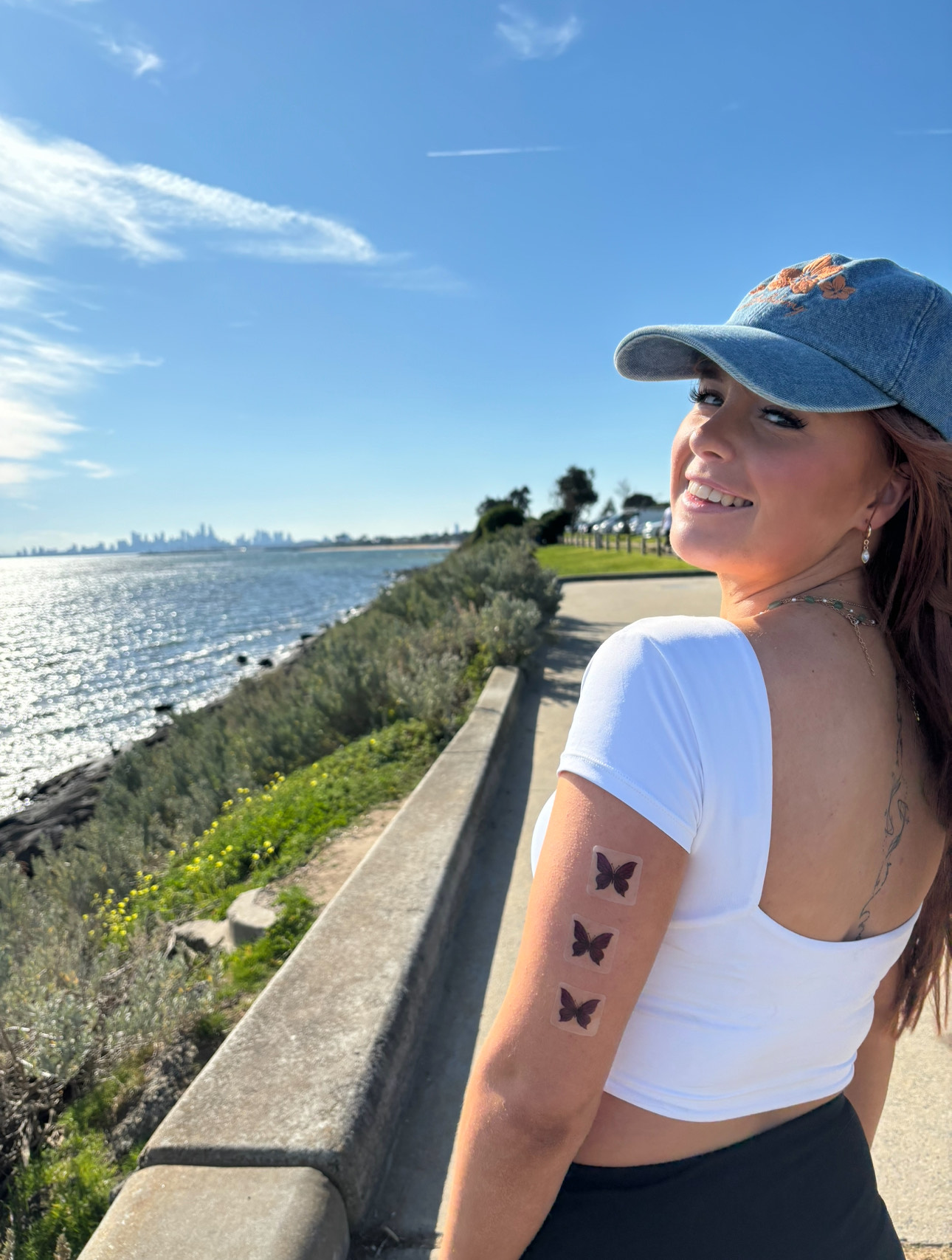 Smiling person with butterfly tattoos on arm, standing by a waterfront path.