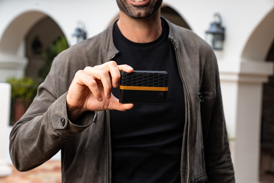 A man in a gray jacket holds a black wallet with a tan stripe across the middle.
