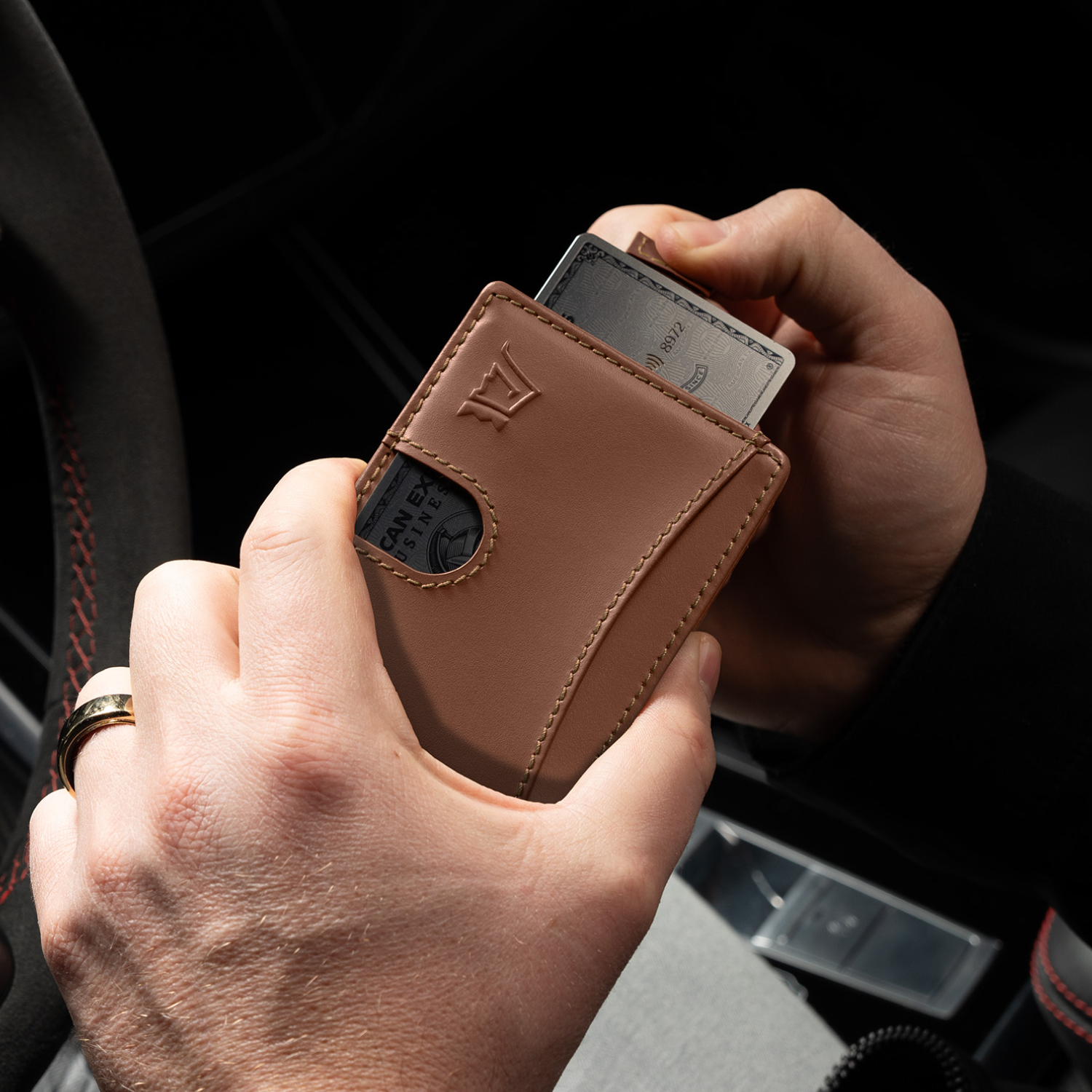 Hands holding a brown wallet with a credit card inside, near a car steering wheel.