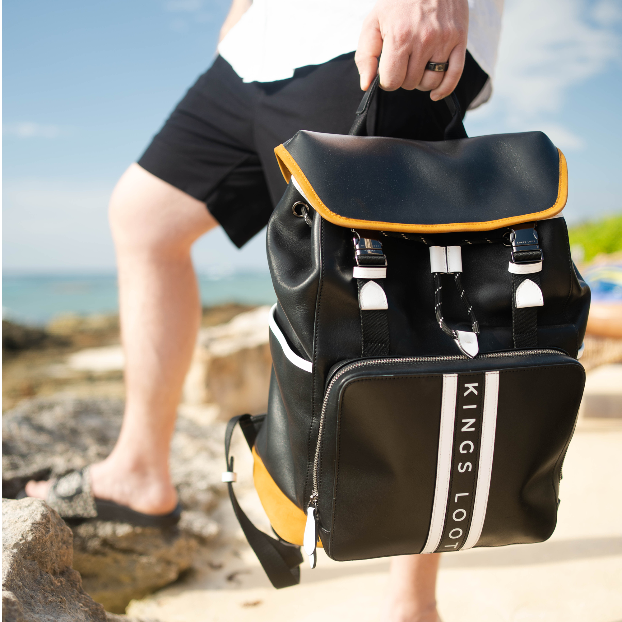 Person holding a black backpack with 'Kings Loot' text, standing on a rocky beach.
